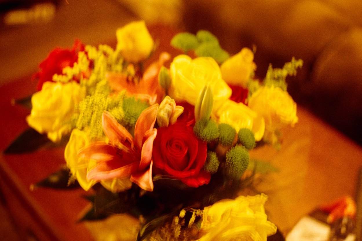 A vibrant floral bouquet featuring bright yellow roses, a prominent red rose, and an orange lily, accented with small green button chrysanthemums and foliage. The arrangement is bathed in warm golden lighting, creating a soft, nostalgic, and intimate atmosphere against a blurred wooden background.